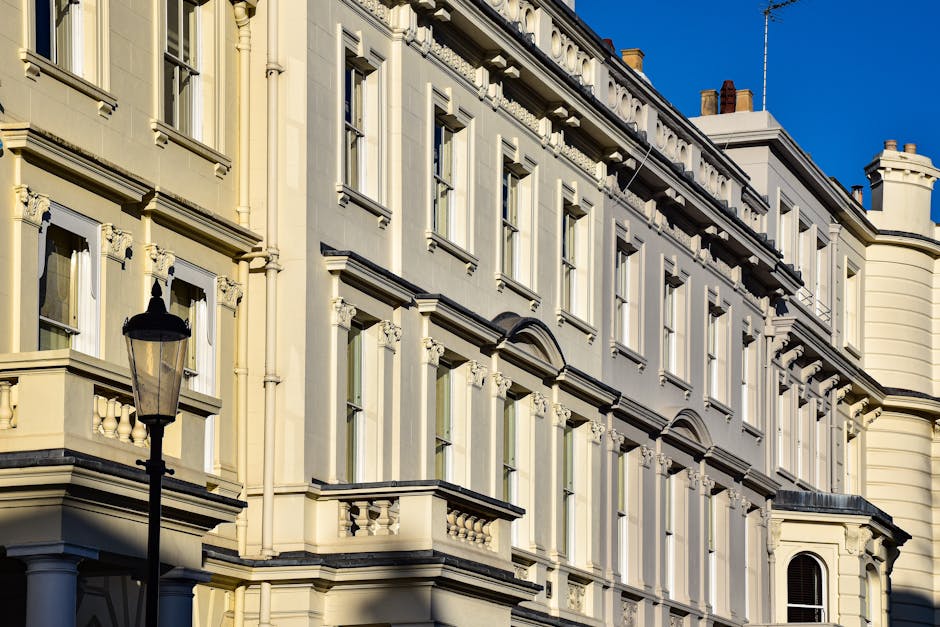 Photograph of a row of elegant, multi-story residential buildings with cream-colored facades, decorative moldings, and black wrought iron balconies, situated along a broad, empty street in Maida Vale.