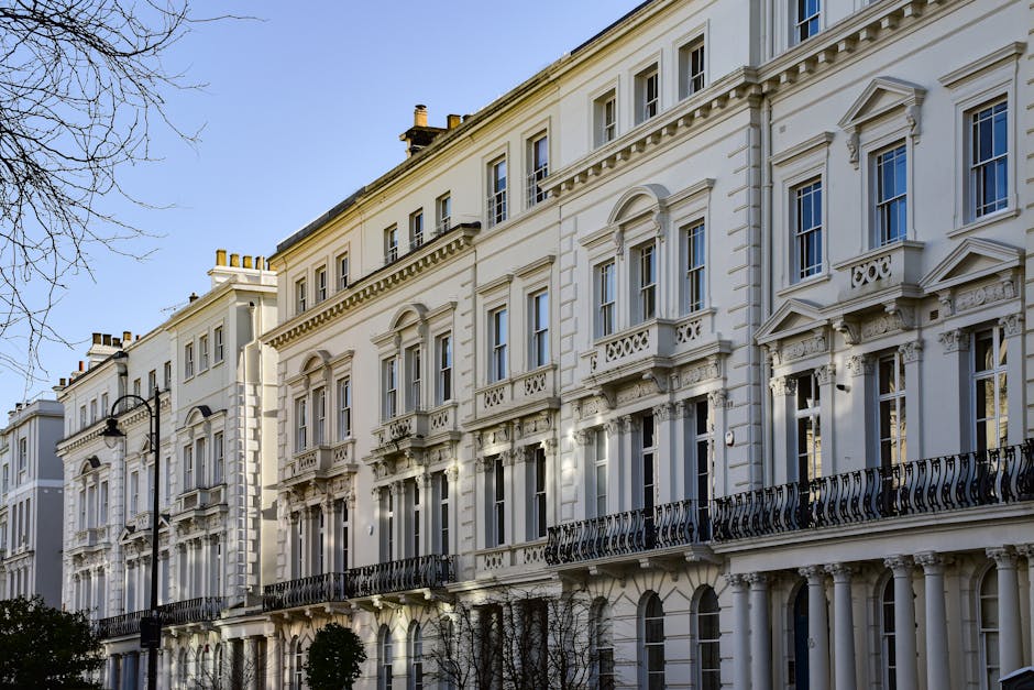 Exterior view of a row of white, Victorian-style terraced townhouses with intricate architectural details, including decorative cornices, arched window frames, and wrought iron balconies, under a clear blue sky. The buildings are illuminated by natural daylight, showcasing their clean facades. In the foreground, a black street lamp is visible, while leafless tree branches partially frame the scene. This image represents well-maintained, historic residential architecture in Maida Vale. Carpet Cleaners Maida Vale provides surface cleaning and deep cleaning services to help preserve the appearance and hygiene of such properties.