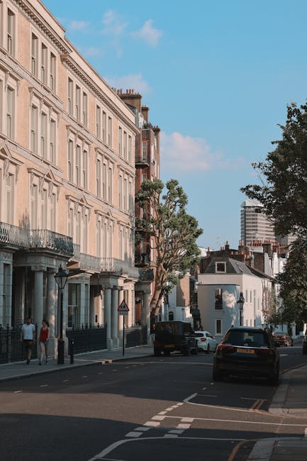 The image depicts a quiet street scene in Maida Vale with a row of classical-style residential buildings featuring cream-colored facades, large windows, and wrought-iron balconies. The street is lined with mature trees providing greenery and shade. Parked cars, including a black vehicle and a white van, are visible along the curb. Pedestrians are walking on the sidewalk, and the sky above is bright with some scattered clouds. The overall scene conveys a clean, well-maintained urban environment typical of Maida Vale, highlighting the area's historic architecture and peaceful atmosphere, which would be complemented by professional domestic cleaning services like those offered by Carpet Cleaners Maida Vale for end of tenancy cleaning tasks.