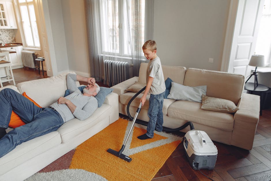 Close-up image of a black vacuum cleaner head cleaning a traditional patterned area rug with floral motifs in soft pastel colors. The vacuum is being operated by a person whose lower legs and white sneakers are visible in the background, standing on a wooden floor partially covered by the rug. The room is well-lit, showcasing the detailed design and clean surface of the rug, demonstrating surface cleaning and deep cleaning processes. The scene highlights home maintenance and hygiene practices, aligning with services provided by Carpet Cleaners Maida Vale for domestic cleaning and sanitisation.