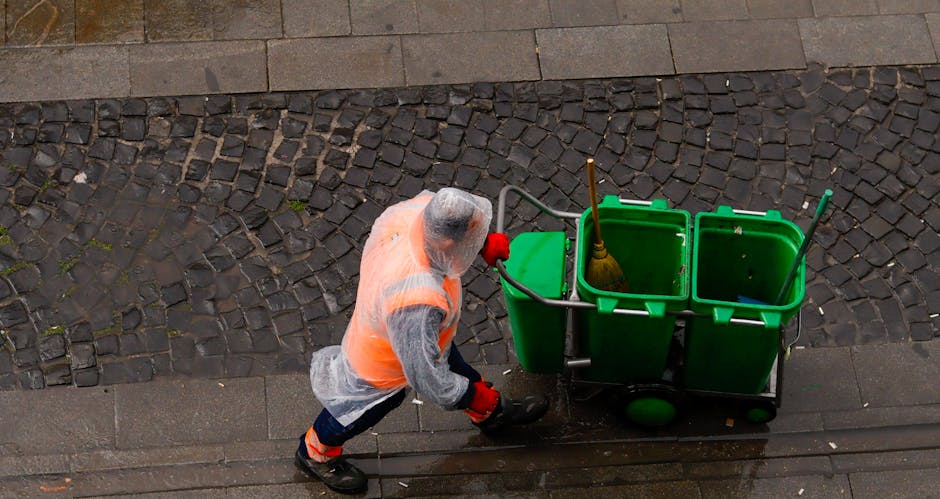 A professional cleaner, dressed in an orange high-visibility vest, grey hoodie, and red gloves, is pushing a green cart containing three open green bins, each with cleaning tools such as a yellow broom and mop. The cleaner is on a sidewalk with a combination of old cobblestone and smooth concrete surfaces, and a wooden tactile paving strip is visible at the edge of the concrete. The scene is illuminated with natural daylight, highlighting the cleanliness of the area. This image relates to surface cleaning and waste management practices, aligning with the Maida Vale council rules for waste from professional cleaners, as detailed by Carpet Cleaners Maida Vale at https://carpetcleanersmaidavale.co.uk for domestic and commercial cleaning services.