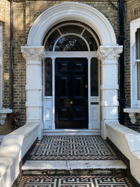 The image depicts the entrance door of a Victorian-style property on Maida Vale W9, framed closely by ornate white stone architraves and columns. Above the black wooden front door, which features a brass doorknob and panel details, there is an semi-circular fanlight window with the number '32' displayed clearly within. The doorstep features patterned black and white mosaic tiling, contrasting with the surrounding brickwork of the building’s facade. White painted brick walls extend on either side of the entrance, and part of a drainpipe is visible on the right. The area is well-lit, with natural daylight highlighting the clean, polished appearance of the surfaces and architectural details, exemplifying the professional surface cleaning and maintenance standards promoted by Carpet Cleaners Maida Vale for Victorian floors and related external features.